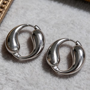 May include: A pair of polished silver hoop earrings. Each earring features a teardrop shape that curves to form a full circle. The earrings are presented on a grey surface.