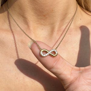 May include: A gold-toned necklace featuring an infinity symbol pendant. One side of the infinity loop is smooth and polished, while the other side is encrusted with small, clear, sparkling stones that catch the light. The pendant hangs from a delicate chain.