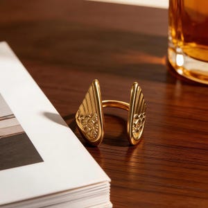 Może przedstawiać: Złoty pierścionek o unikalnym wzorze. Pierścionek posiada dwa elementy w kształcie skrzydeł ze skomplikowanymi detalami, połączone obrączką. Leży na ciemnej drewnianej powierzchni obok czasopisma i szklanki.