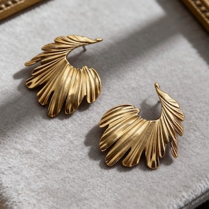 May include: A pair of gold-coloured earrings with a leaf-like design. Each earring features a curved, fan-shaped structure with detailed, ridged textures, creating a natural, organic aesthetic. The earrings are displayed on a soft, grey surface.
