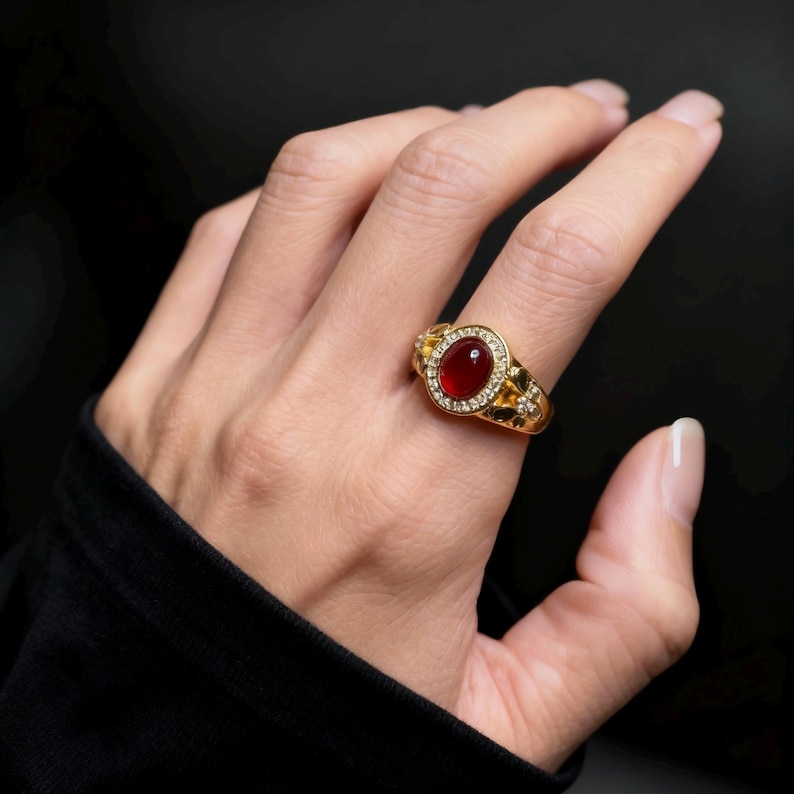 May include: A gold-colored ring featuring a large, oval, red gemstone at its center, surrounded by smaller, clear stones. The ring has an ornate design with decorative elements on the band. The ring is on a person's finger against a dark background.