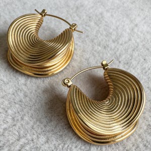 May include: A pair of gold-coloured hoop earrings. Each earring has a layered, semi-circular design with concentric ridges. The earrings have a classic hinged closure and are displayed on a neutral grey surface.