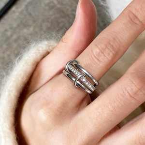 May include: A silver ring with a distinctive design. The ring has three bands, one of which is set with small, clear stones. The bands are interwoven, creating a modern and stylish appearance. The ring is shown on a finger.