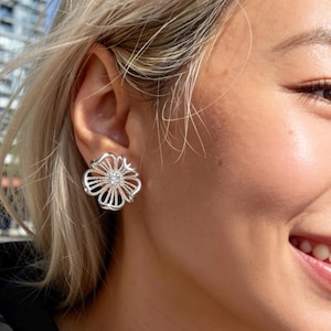 May include: A silver flower-shaped earring with a central clear stone. The earring features an openwork design with multiple petals and a sparkling centre, reflecting light.