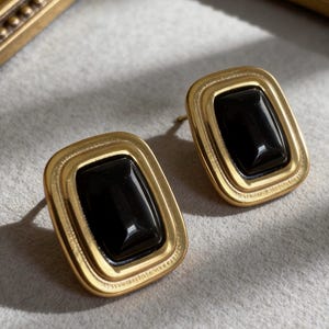 May include: A pair of gold-toned stud earrings with a rectangular black stone center. The earrings have a double-layered frame design. The earrings are displayed on a light gray surface.