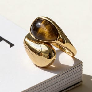 May include: A gold ring featuring a large, oval tiger's eye gemstone. The ring has a distinctive, open design with a smooth, polished finish. The stone displays brown and gold hues.