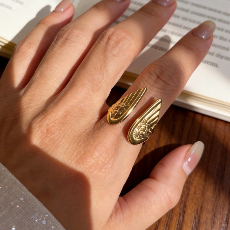 Może przedstawiać: Złoty pierścionek z motywem skrzydeł. Pierścionek posiada dwa otwarte skrzydła, każde ze szczeg&oacute;łowymi wzorami pi&oacute;r i kwiat&oacute;w. Pierścionek jest noszony na palcu, ze skrzydłami rozciągającymi się na zewnątrz.