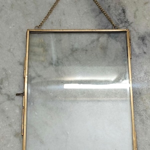 Pode incluir: Uma moldura retangular com uma borda fina de metal em tom de latão e dois painéis de vidro transparente. A moldura possui um fecho lateral articulado e uma delicada corrente de metal a condizer no topo para pendurar.