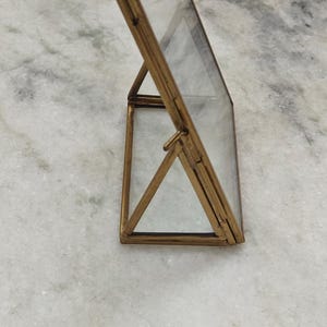 May include: A vertical rectangular picture frame constructed with a thin brass metal frame and clear glass panels. The frame features a hinged design with a triangular support stand that allows it to stand upright on a flat surface.