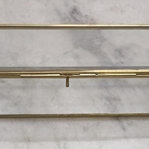 Puede incluir: Una caja expositora rectangular construida con paneles de cristal transparente y un marco de metal en tono dorado. La caja cuenta con una tapa abatible con un fino mecanismo de cierre metálico centrado en el borde frontal.