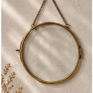 May include: A circular floating frame with a metallic brass finish, featuring two clear glass panes held within a thin metal border. The frame includes a hinged side closure and is suspended by a matching brass chain attached to a small wall hook.