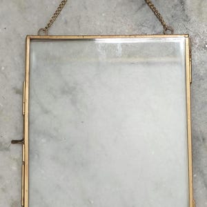 May include: A rectangular floating picture frame with a thin gold-toned metal border and two clear glass panes. The frame features a hinged side closure and a delicate gold-toned metal chain attached to the top for hanging display purposes.