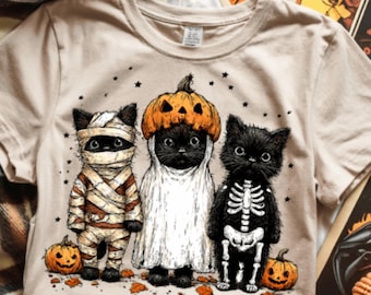 May include: Beige t-shirt featuring a Halloween-themed graphic of three black cats dressed as a mummy, ghost, and skeleton. Each cat has a pumpkin and is surrounded by stars and leaves. The shirt is made of a soft material.