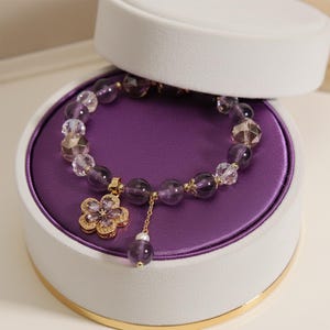 Op de afbeelding: Een paarse kralenarmband met een gouden bloemvormige bedel en een bungelende amethist kraal, gepresenteerd in een wit-gouden sieradendoos met een paars interieur. De armband heeft een mix van ronde en facet geslepen kralen.