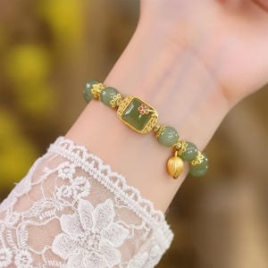 Puede incluir: Una pulsera con cuentas de jade verde y detalles dorados. Presenta una pieza central cuadrada de jade con un pequeño diseño de flor roja y un dije de loto dorado. La pulsera se lleva en la muñeca.