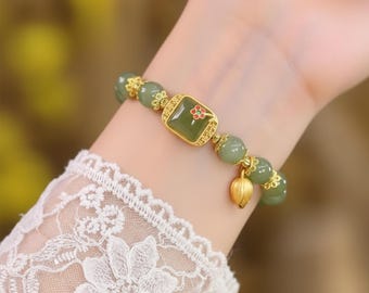 May include: A beaded bracelet featuring green jade beads and gold-colored accents. The bracelet includes a square jade centerpiece with a small red flower design and a gold lotus charm. The bracelet is worn on a wrist.