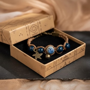 Puede incluir: Una pulsera artesanal con cuentas azules, una cuenta central azul y blanca, y un dije de estrella de mar de bronce. La pulsera se presenta en una caja de cartón decorativa con un patrón geométrico.