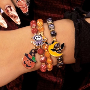 Könnte beinhalten: Drei Armbänder mit Halloween-Anhängern. Die Anhänger umfassen einen Kürbis, einen Geist und eine Fledermaus mit einem Halbmond. Die Armbänder haben rote, orangefarbene und schwarze Perlen.