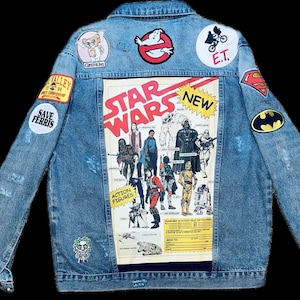 Puede incluir: Chaqueta vaquera azul claro con varios parches de cultura pop, incluyendo "Gremlins", "Ghostbusters", "E.T.", "Superman" y "Batman". La parte trasera presenta un gran póster de "Star Wars" con figuras de acción. La chaqueta tiene un aspecto desgastado.