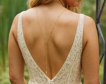 Könnte beinhalten: Eine goldfarbene Rückenkette mit zarter Kette und kleinen Perlenakzenten. Die Kette verläuft über den Rücken und bildet ein Y. Das Model trägt ein weißes Brautkleid mit tiefem Rückenausschnitt und floralem Spitzenmuster.