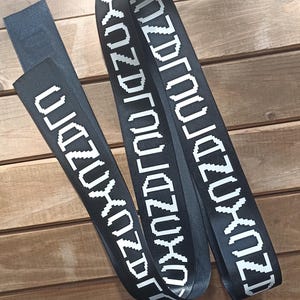 Op de afbeelding: Twee zwarte stoffen banden met een herhalend wit gepixeld tekstpatroon dat "KUZDJOJAZUXO" spelt. De banden zijn gemaakt van een glad, duurzaam materiaal en tonen de witte letters duidelijk tegen de donkere achtergrond in een consistent lettertype.