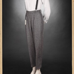 Puede incluir: Pantal&oacute;n de lana gris de cintura alta y pierna ancha. El pantal&oacute;n se sujeta con tirantes negros. El pantal&oacute;n se combina con una camisa blanca abotonada con finas rayas azules. La modelo lleva el atuendo con una expresi&oacute;n neutral.