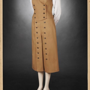 K&ouml;nnte beinhalten: Beigefarbenes Outfit im Vintage-Stil auf einer Schaufensterpuppe. Das Ensemble umfasst eine doppelreihige Weste, eine hoch taillierte Culotte mit Knopfdetails, ein wei&szlig;es Hemd mit Kragen und eine gemusterte Krawatte. Das Outfit wird vor dunklem Hintergrund pr&auml;sentiert.