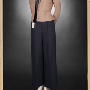 Puede incluir: Una chaqueta de color beige con cuello ancho y dos bolsillos de solapa. La chaqueta se lleva sobre una camisa blanca con una corbata de estampado negro y unos pantalones negros. Los pantalones tienen una pierna ancha y est&aacute;n plisados en la cintura. La modelo lleva zapatos negros.