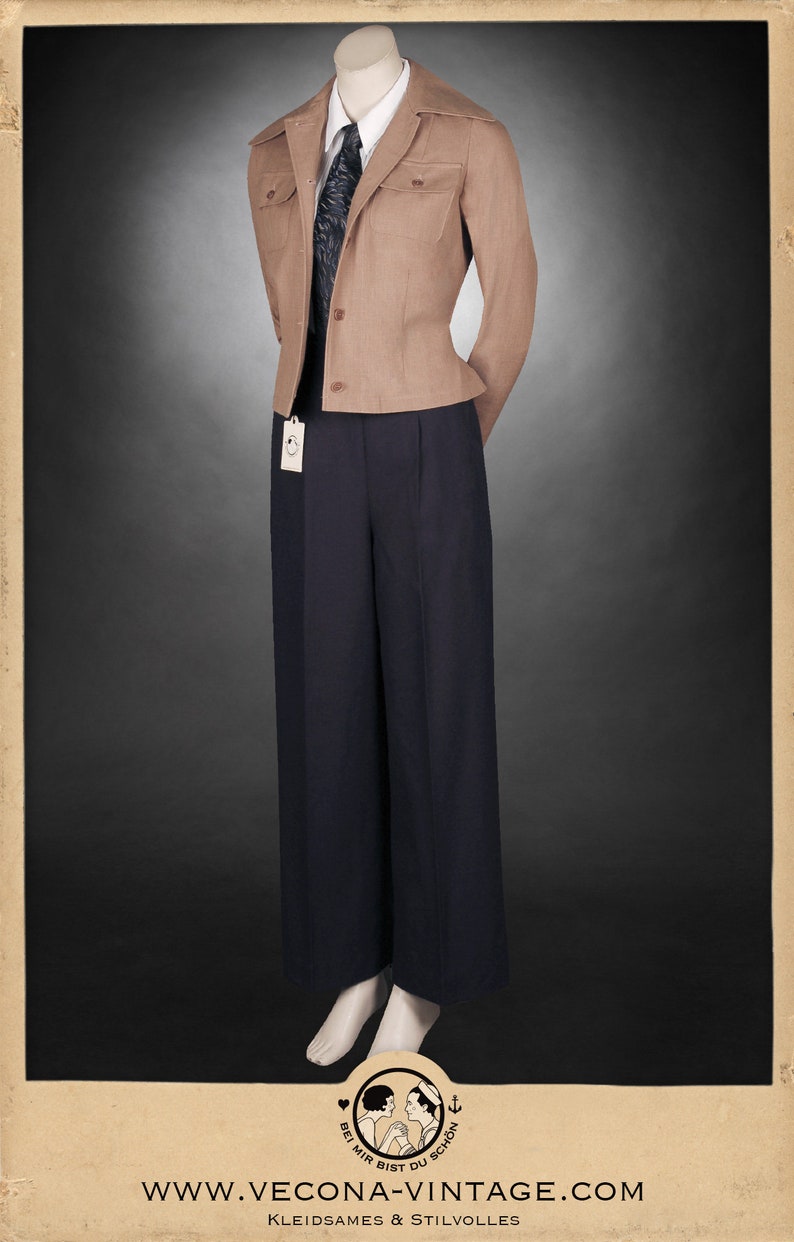 Puede incluir: Una chaqueta de color beige con cuello ancho y dos bolsillos de solapa. La chaqueta se lleva sobre una camisa blanca con una corbata de estampado negro y unos pantalones negros. Los pantalones tienen una pierna ancha y est&aacute;n plisados en la cintura. La modelo lleva zapatos negros.