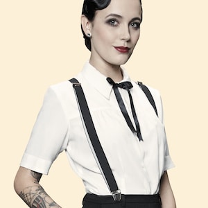 May include: A woman wearing a white button-down shirt with black suspenders and a black bow tie. She has dark hair styled in a vintage updo and is wearing black pants.