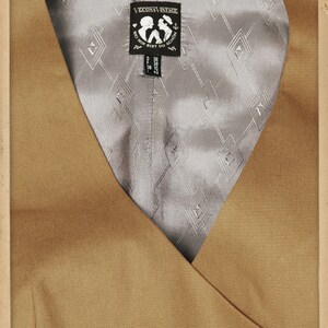 K&ouml;nnte beinhalten: Beige Weste mit V-Ausschnitt. Das Innenfutter ist ein hellgrauer Stoff mit Rautenmuster. Ein schwarzes Etikett mit dem Text "VECONA VINTAGE" ist sichtbar. Die Website-Adresse lautet www.vecona-vintage.com.