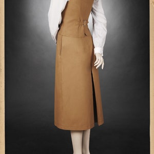 K&ouml;nnte beinhalten: Ein beigefarbener Vintage-Rockanzug mit einem langen Rock mit Schlitz und einer passenden Weste. Die Weste hat eine Schnalle am R&uuml;cken. Darunter wird ein wei&szlig;es gestreiftes Hemd getragen. Das Outfit wird auf einer Schaufensterpuppe pr&auml;sentiert.