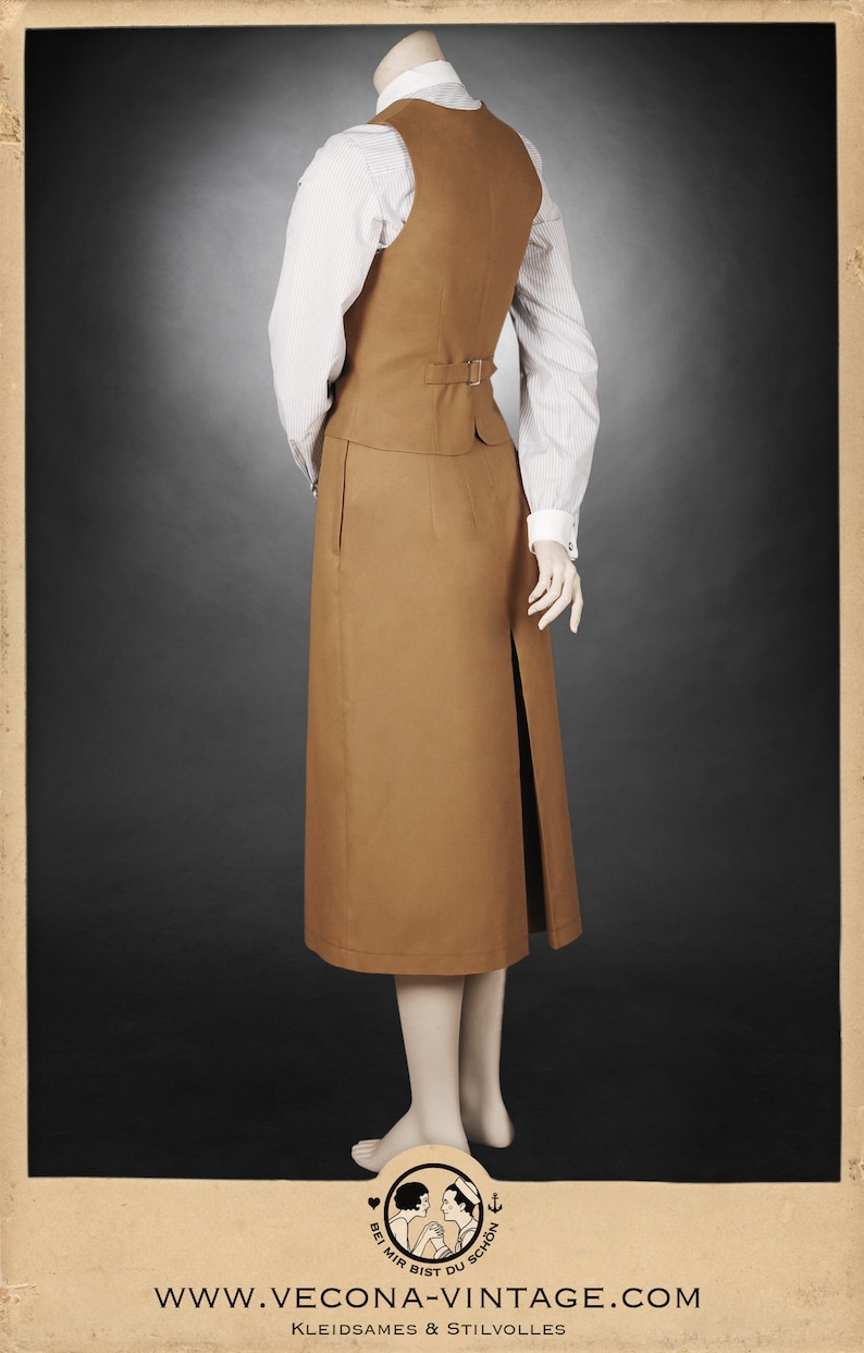 K&ouml;nnte beinhalten: Ein beigefarbener Vintage-Rockanzug mit einem langen Rock mit Schlitz und einer passenden Weste. Die Weste hat eine Schnalle am R&uuml;cken. Darunter wird ein wei&szlig;es gestreiftes Hemd getragen. Das Outfit wird auf einer Schaufensterpuppe pr&auml;sentiert.
