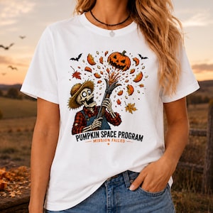 May include: A white t-shirt featuring a graphic of a skeleton wearing a straw hat and red plaid shirt, launching a jack-o'-lantern from a cannon. The design includes flying pumpkin pieces, autumn leaves, and the text PUMPKIN SPACE PROGRAM MISSION FAILED.