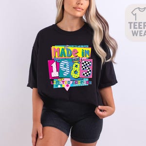 May include: Black t-shirt with a colorful graphic that reads "MADE IN 1985 LIMITED EDITION." The design features the numbers 1, 9, 8, and 5 in various bright colors and patterns, including pink, blue, yellow, and checkered. The shirt is a casual style.