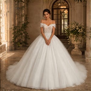 Könnte beinhalten: Ein weißes Ballkleid-Brautkleid mit schulterfreiem Ausschnitt, einem eng anliegenden Oberteil mit aufwendigen Spitzenverzierungen und einem voluminösen, bodenlangen Tüllrock, der sich ab der Taille ausstellt und eine volle, dramatische Silhouette bildet.