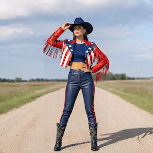 Pode incluir: Uma mulher vestindo uma jaqueta de couro vermelha, branca e azul com franjas, um top curto azul marinho e calças de couro azul marinho com listras vermelhas. Ela também está usando um chapéu de cowboy azul marinho e botas de cowboy pretas. A roupa é usada em uma estrada de terra.
