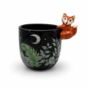 May include: A black ceramic mug featuring a painted design of green ferns, leaves, and a white crescent moon with small stars. A three-dimensional orange and white fox figure is attached to the upper rim, serving as a decorative handle element.