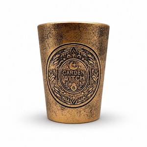 Könnte beinhalten: Ein konisch zulaufendes zylindrisches Gefäß mit einer abgenutzten, metallisch bronzefarbenen Oberfläche. Die Vorderseite zeigt ein kreisförmiges geprägtes Emblem mit dem Text GARDEN WITCH, umgeben von Blattmotiven, einer Mondsichel und kleinen Sternen.