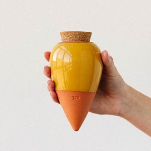 Può includere: Un vaso di ceramica giallo con un tappo di sughero, tenuto in mano. Il vaso ha una base a forma di cono color terracotta. La superficie lucida del vaso riflette la luce. Il vaso è alto circa 15 cm.