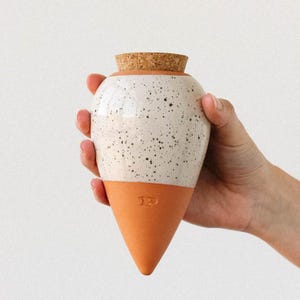 Può includere: Un vaso in ceramica bianca e arancione macchiata con tappo in sughero. Il vaso ha una parte superiore arrotondata e una base a forma di cono. Il vaso è tenuto in mano su sfondo bianco.