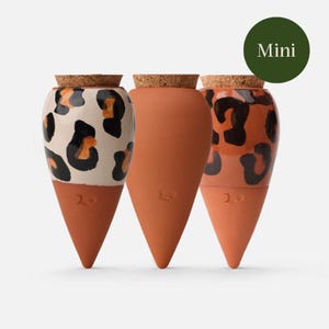 Peut inclure: Trois mini-pots en terre cuite avec bouchons en liège. L'un des pots a une base blanche avec un imprimé léopard noir et orange. Les deux autres sont en terre cuite unie. Le mot "Mini" est dans un cercle vert en haut à droite.