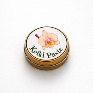 Könnte beinhalten: Kleine, runde, goldfarbene Dose mit einem weißen Etikett, das eine Aquarellillustration einer hellrosa Orchideenblüte zeigt. Der Text "Keiki Paste" ist darunter in Grün gedruckt. Die Dose wird wahrscheinlich für ein Pflanzenpflegeprodukt verwendet.