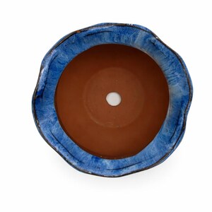 May include: Overhead view of a ceramic planter with a wavy, irregular rim. The rim is glazed in shades of blue, contrasting with the unglazed, reddish-brown interior. A drainage hole is visible at the bottom. The planter is designed for indoor or outdoor use.