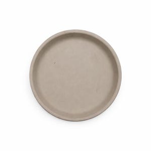 May include: A round, light beige concrete tray. The tray has a shallow, recessed center and a raised, rounded edge. The neutral color and simple design make it suitable for various uses, such as a decorative piece or a functional item.