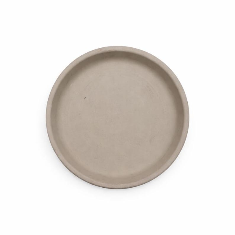 May include: A round, light beige concrete tray. The tray has a shallow, recessed center and a raised, rounded edge. The neutral color and simple design make it suitable for various uses, such as a decorative piece or a functional item.