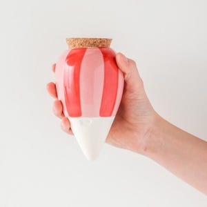 Może przedstawiać: Różowo-czerwony, pasiasty ceramiczny wazon z korkiem. Wazon ma białą, stożkową podstawę. Wazon jest trzymany w dłoni osoby na białym tle.