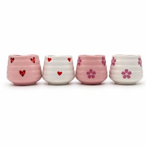Può includere: Quattro tazze da tè in ceramica in fila. Due sono rosa con cuori rossi, una è bianca con cuori rossi e l'ultima è rosa con fiori di ciliegio rosa. Le tazze hanno un design strutturato e scanalato. Ideali per tè o caffè.
