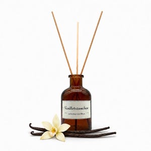 May include: A brown glass bottle reed diffuser with three wooden sticks. The white label features the text Vanilleträumchen and enchanting room diffuser. A single cream-coloured vanilla flower and two dark brown vanilla beans lie at the base.