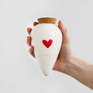 Può includere: Un vaso in ceramica bianco con un cuore rosso dipinto sul davanti. Il vaso ha un tappo di sughero ed è tenuto in mano. Il vaso ha una forma a goccia e una finitura lucida.